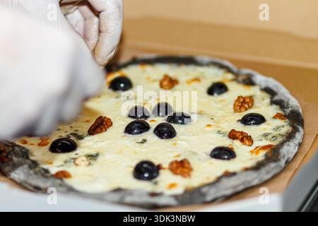Chef hand adding black olives and walnuts to baked cheese pizza on black dough in box. Finishing touch. Stock Photo