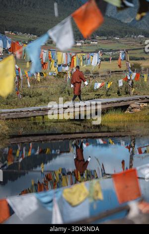 View of a man walking on a wooden bridge over a reflective lake under prayer flags in the valley, Phobjikha Valley, Wangdue Phodrang, Bhutan. Stock Photo