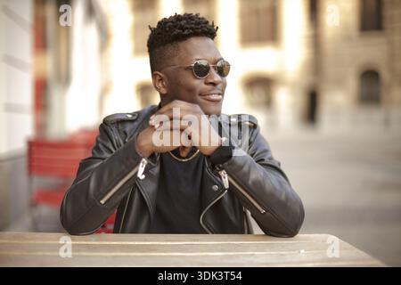 Thoughtful person in a black jacket and sunglasses sitting at an outdoor table of street cafe or restaurant feeling charmed, charming smile, single, Stock Photo