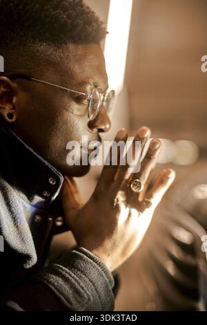 Sideview portrait of confident young man with folded hands, a man in glasses appears deep in thought or prayer with hands clasped, lit warmly, wearing Stock Photo