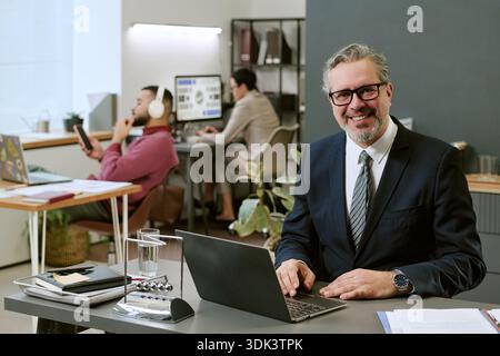 Middle Aged Man Smiling While Working on Laptop in Modern Office Stock Photo