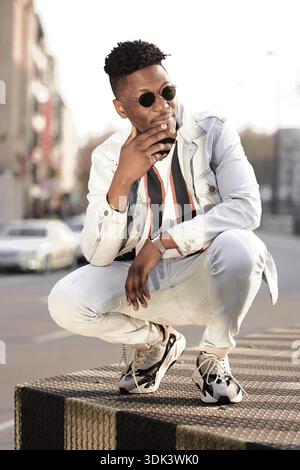 Smitty man stroking his chin in a thoughtful manner of cleverness, crouching on a street wearing a denim jacket and sunglasses, serious, ernest, conte Stock Photo
