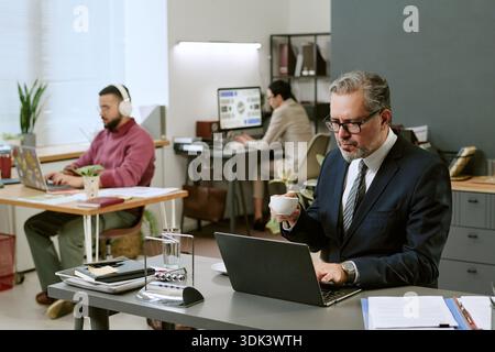 Middle Aged Man Working on Laptop While Drinking Coffee in Office Stock Photo