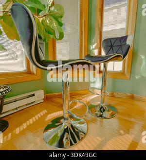 Chrome pedestal chairs decorate this interior alcove kitchen space. Light from windows splashes shadows on plants & furnishings. Floor is wood. Stock Photo