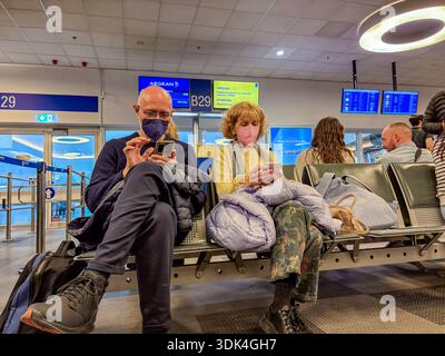 Elderly couple wearing protective face masks, using smartphones while waiting at an airport gate (Gate B29, Aegean Airlines visible). Travel, waiting, Stock Photo