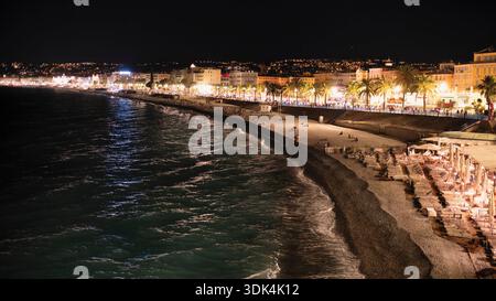 The Beach of Nice at Night with Lounge Chairs and People relaxing with the Promenade des Anglais in the background with Palm Trees and the Old Town, A Stock Photo