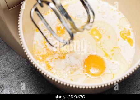 Stand mixer bowl with eggs and milk closeup. Baking ingredients preparation process. Electric kitchen appliance mixing dough. Home cooking and pastry. Stock Photo