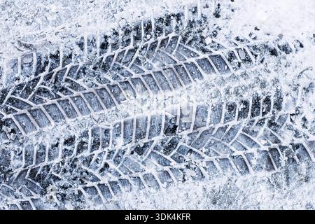 Car tire tracks in snow closeup. Winter road surface texture. Icy frozen ground with vehicle tread marks. Cold season transport background. Stock Photo