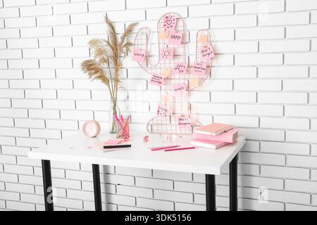 Pinboard with sticky notes, vase and stationery on table near white brick wall Stock Photo