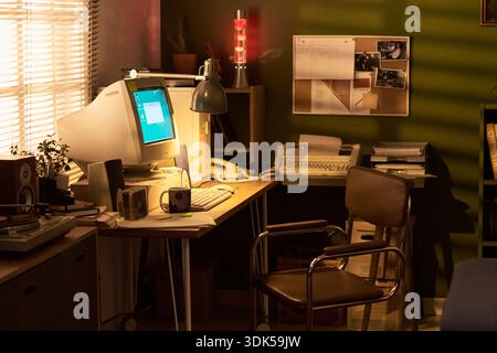 Vintage home office scene featuring empty chair positioned at wooden desk with old CRT computer monitor, keyboard, desk lamp, telephone, lava lamp, coffee mugs, and corkboard on green wall Stock Photo