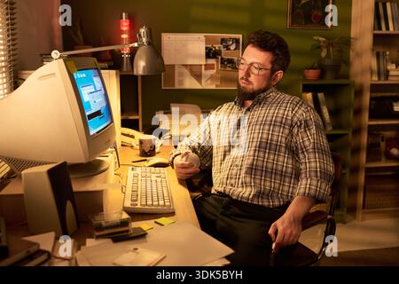 Caucasian man sitting at desk using vintage desktop computer, holding baseball in hand, wearing glasses, looking at monitor in home office environment Stock Photo