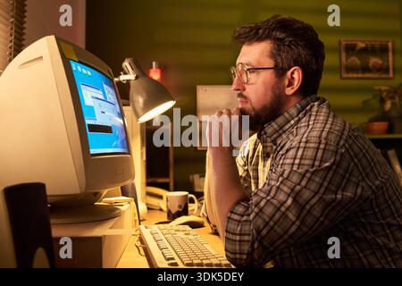 Caucasian man with beard and glasses sitting at desk using vintage CRT computer monitor, resting chin on hand and focusing on screen in office setting Stock Photo