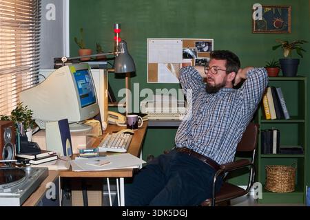 Caucasian man with beard leaning back in chair relaxing at vintage office desk using retro computer surrounded by paperwork, books, and plants Stock Photo