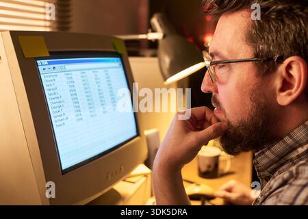 Caucasian man wearing glasses working at desk, analyzing spreadsheet data on computer monitor, resting chin on hand, focused on screen in office setting Stock Photo