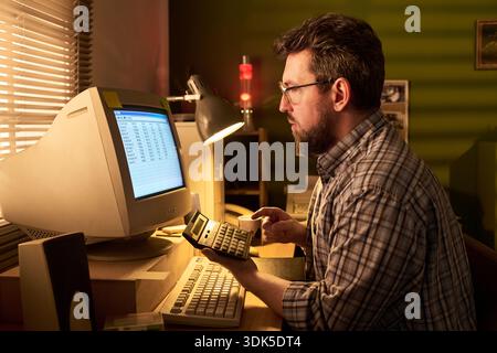 Caucasian man working at desk using vintage computer and calculator, analyzing data on monitor, sitting in office with focused expression, holding calculator in hand Stock Photo