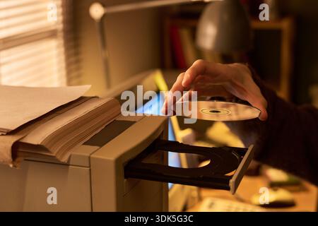 Caucasian woman inserting compact disc into computer disc drive, hand visible holding CD above open tray, stack of documents on system unit, office workspace in background Stock Photo