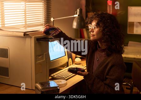 Caucasian woman inserting compact disc into vintage desktop computer, holding stack of CDs in hand, sitting at desk with retro monitor and keyboard, wearing eyeglasses Stock Photo