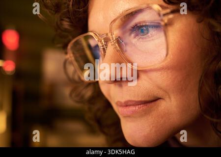 Portrait of Caucasian woman wearing eyeglasses focusing intently while working on computer screen, close up showing facial features and curly hair in soft lighting Stock Photo