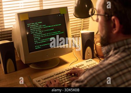 Caucasian middle aged man typing on vintage computer with green text on CRT monitor, sitting at wooden desk with speakers and blinds in background, focusing on programming code Stock Photo