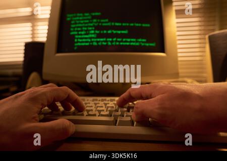 Man typing on computer keyboard with green code displayed on monitor, hands in foreground, working in dimly lit office environment with blinds in background Stock Photo