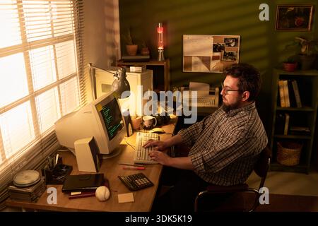 Caucasian man sitting at desk working on vintage computer in retro office setting, typing on keyboard with focused expression, surrounded by old technology equipment Stock Photo
