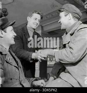 Slaviansk, Ukrainian SSR, Soviet Union - 1960s: Three cheerful male workers shake hands in the cultural club of the Slaviansk Ceramic Combine. Two employees in work jackets and caps greet each other while a young colleague looks on with a smile. The interior features decorated ceilings and columns. This black and white archival image captures the atmosphere of male friendship and solidarity in the peaceful Donbas. The vintage shot symbolizes the social life and camaraderie of the Soviet proletariat during the mid-20th century. Stock Photo
