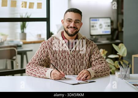 Portrait of Young Adult Man Smiling While Writing at Desk Stock Photo