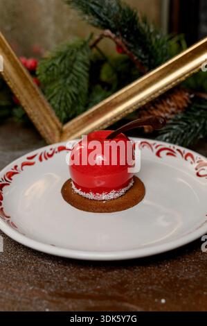 Red apple shaped cake with glossy icing on a white plate.A bright red mousse cake on a plate in a cozy New Year's interior with pine needles Stock Photo