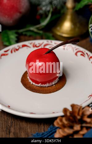 Red apple shaped cake with glossy icing on a white plate.A bright red mousse cake on a plate in a cozy New Year's interior with pine needles Stock Photo