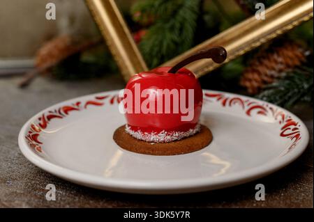 Red apple shaped cake with glossy icing on a white plate.A bright red mousse cake on a plate in a cozy New Year's interior with pine needles Stock Photo