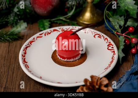 Red apple shaped cake with glossy icing on a white plate.A bright red mousse cake on a plate in a cozy New Year's interior with pine needles Stock Photo
