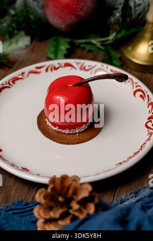Red apple shaped cake with glossy icing on a white plate.A bright red mousse cake on a plate in a cozy New Year's interior with pine needles Stock Photo