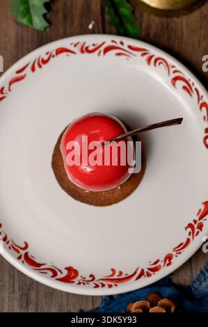 Red apple shaped cake with glossy icing on a white plate.A bright red mousse cake on a plate in a cozy New Year's interior with pine needles Stock Photo