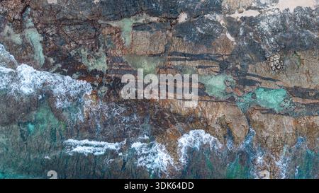 Aerial view of rocky shoreline with turquoise sea, waves breaking into white foam, layered stone textures and natural abstract pattern. Stock Photo