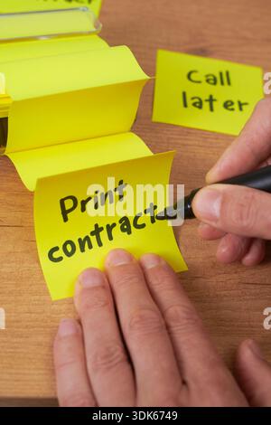 a close-up of a man writing print contract on a yellow sticky note placed on a wooden desk, with other notes and a marker holder visible nearby Stock Photo