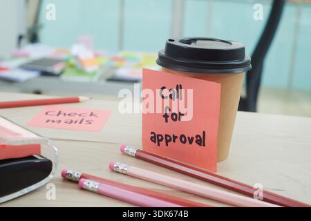 a disposable coffee cup on an office desk with a pink sticky note reading call for approval attached to it, surrounded by pencils, notes, and everyday Stock Photo
