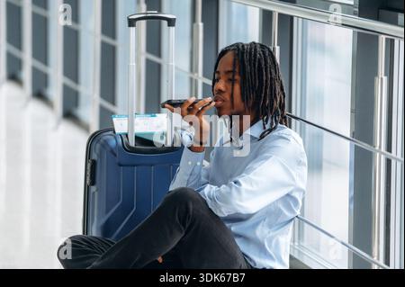 Sitting on the floor with phone in hand. Male tourist is in the airport corridor. Stock Photo