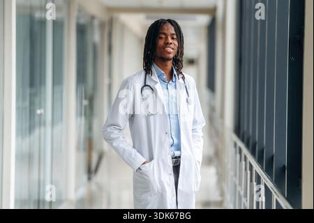 Positive facial expression, standing. Young male black doctor is in the corridor. Stock Photo