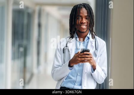 Cheerful, with phone. Young male black doctor is in the corridor. Stock Photo