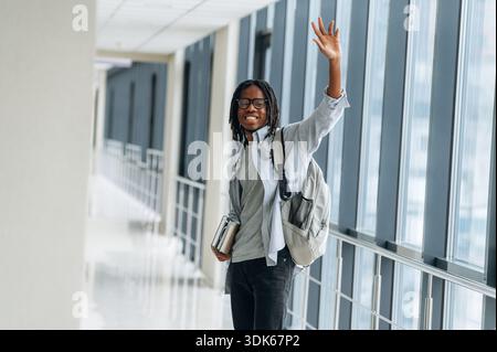 Bye hand gesture, smiling. Student with backpack is in the corridor. Stock Photo