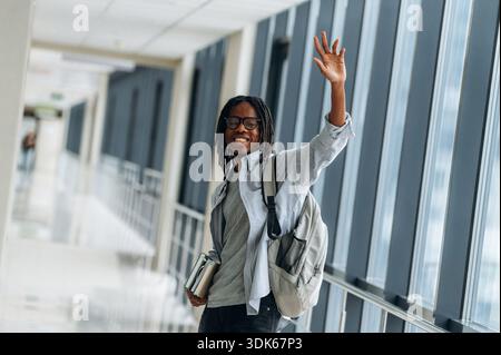 Bye hand gesture, smiling. Student with backpack is in the corridor. Stock Photo