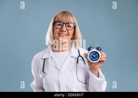Clock in hand, smiling. Senior woman doctor is against background. Stock Photo
