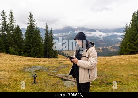 Man using drone controller in mountain meadow. Man in hooded jacket operating a drone controller in an open mountain field, surrounded by pine trees Stock Photo