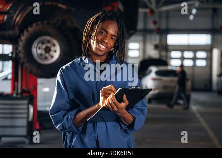 Taking notes into notepad. Male mechanic is with car in garage. Stock Photo
