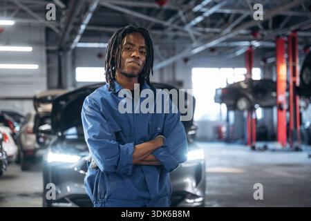 Friendly male mechanic is with car in garage, standing with arms crossed. Stock Photo