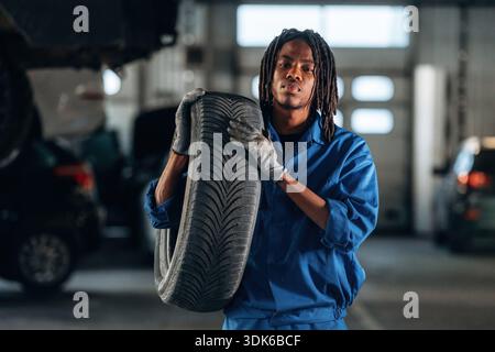 Heavy tire, holding it, changing. Male mechanic is with car in garage. Stock Photo