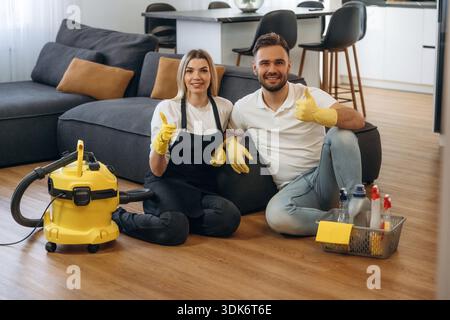 Staff sitting on floor, smiling, rest. Two cleaning service workers are in domestic room. Stock Photo
