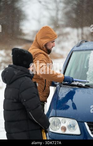 A man in a brown jacket clears snow from a blue car's windshield using an ice scraper. A child in a black jacket observes nearby on a snowy day. Stock Photo