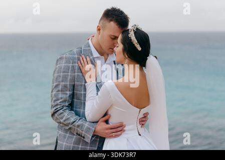 A bride and groom share an intimate moment during their wedding ceremony by the water, with the groom in a plaid suit and the bride in a white gown Stock Photo