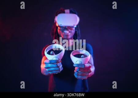 Positive, smiling and holding controllers. African American guy is with virtual reality glasses in neon room. Stock Photo